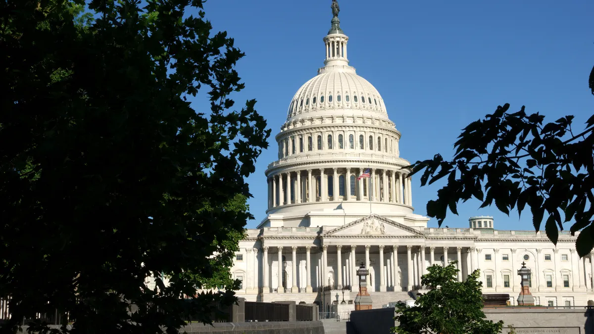 A view of the front of the U.S. Capitol building framed by green-leaf trees against a clear blue sky.