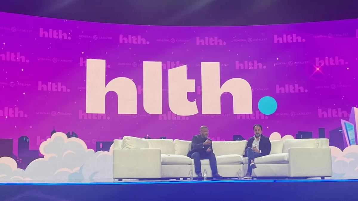 Hemant Taneja and Marc Harrison sit on a white couch on a large stage at the HLTH conference.