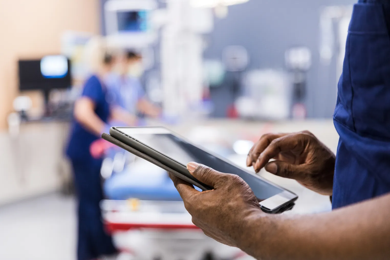 A healthcare worker in blue scrubs holds a tablet in a clinical setting