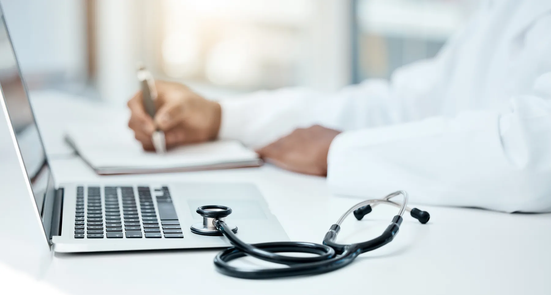 Laptop, stethoscope and doctor writing in notebook for research planning or medical tech innovation in hospital office.