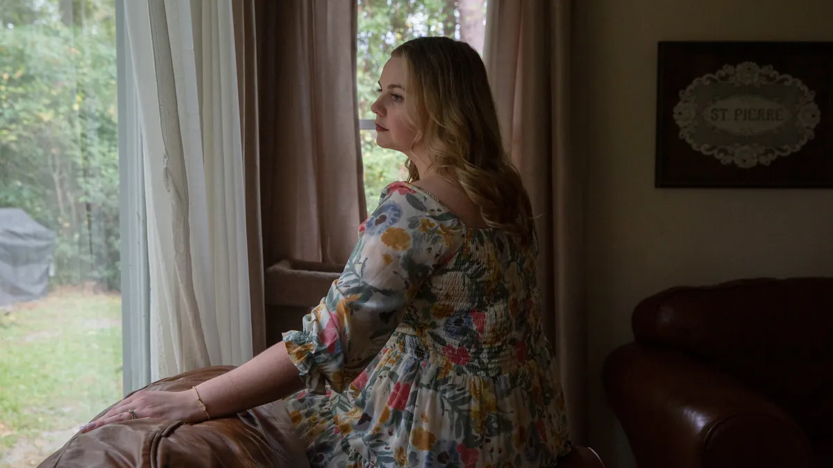 A person sits on the edge of a couch looking out a window in a living room setting.